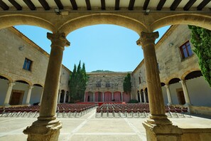 Une cour ouverte, avec des rangées de chaises, des colonnes de pierre et un bâtiment doté de fenêtres en arc de cercle et d’une scène.