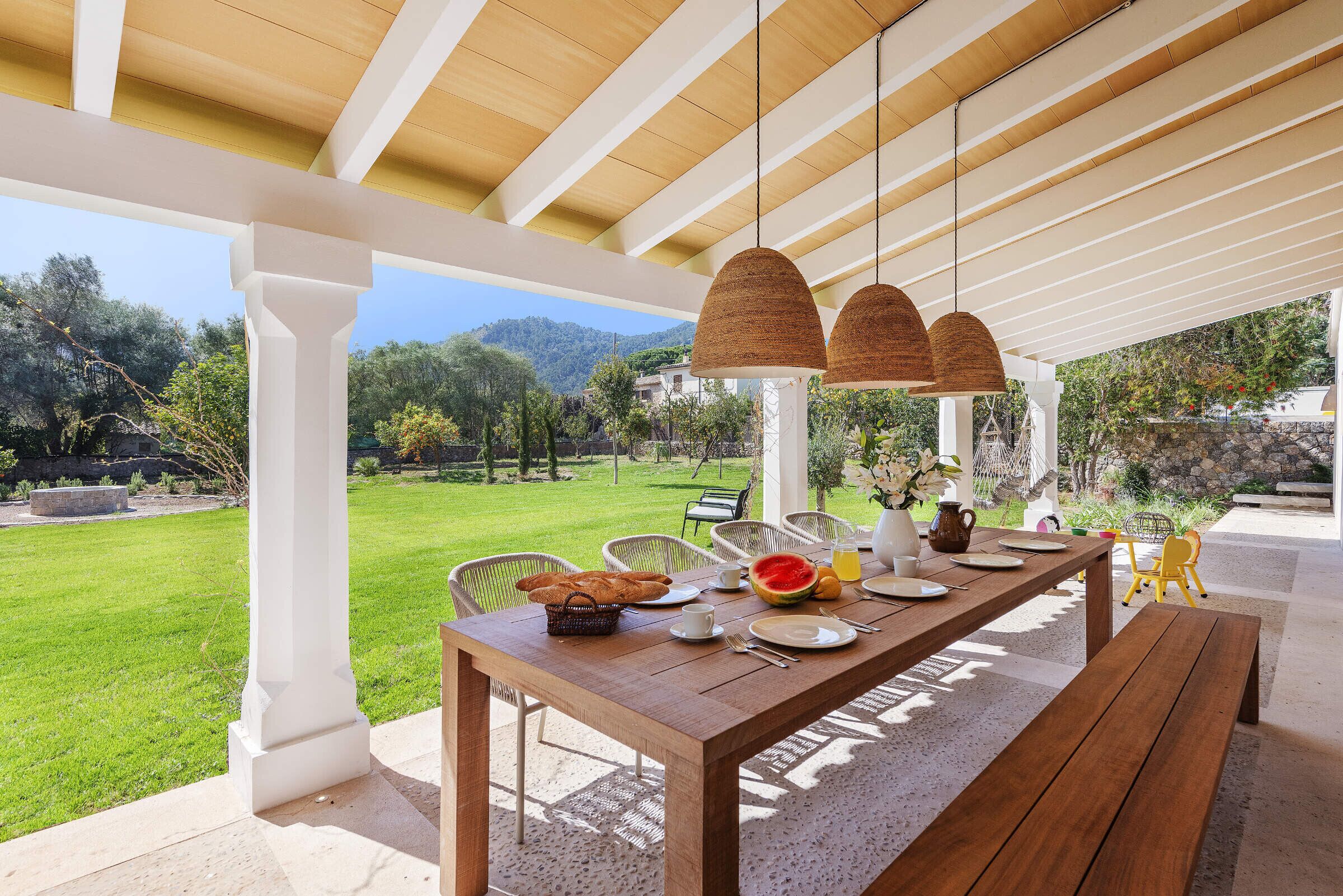 An outdoor dining area with a wooden table, white chairs, and hanging wicker lamps.