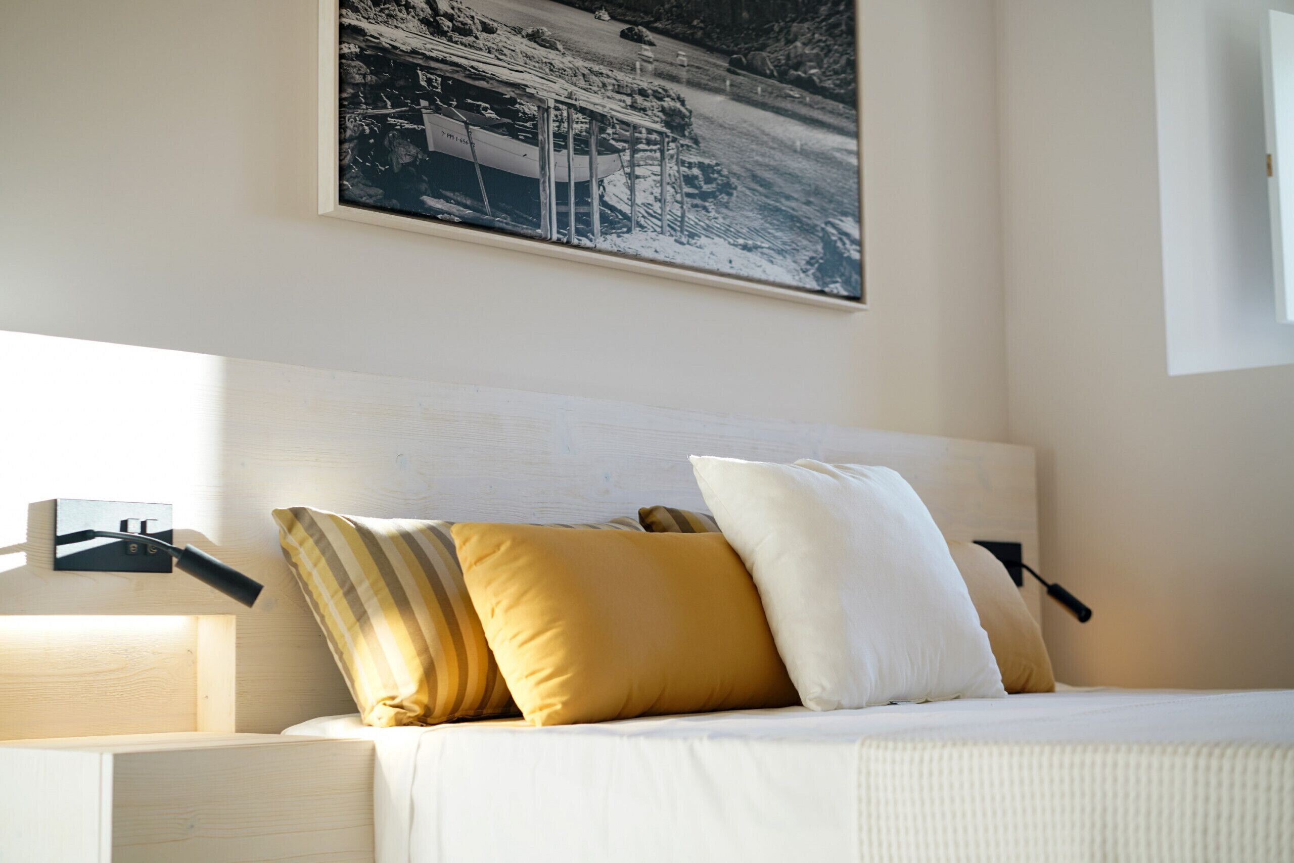A neatly made bed with white and mustard pillows, a wooden headboard, and a black lamp on a bedside table.