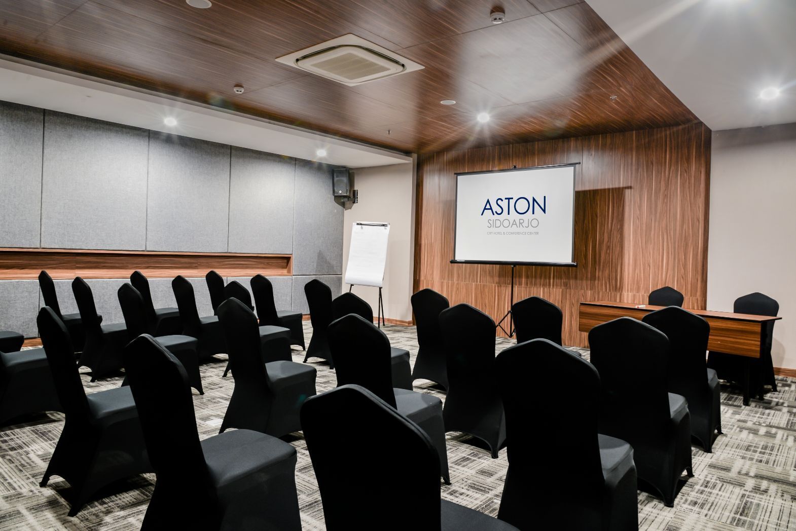 A conference room with rows of chairs, a projector screen displaying "ASTON SIDOARJO CITY HOTEL & CONFERENCE CENTER", and a whiteboard.