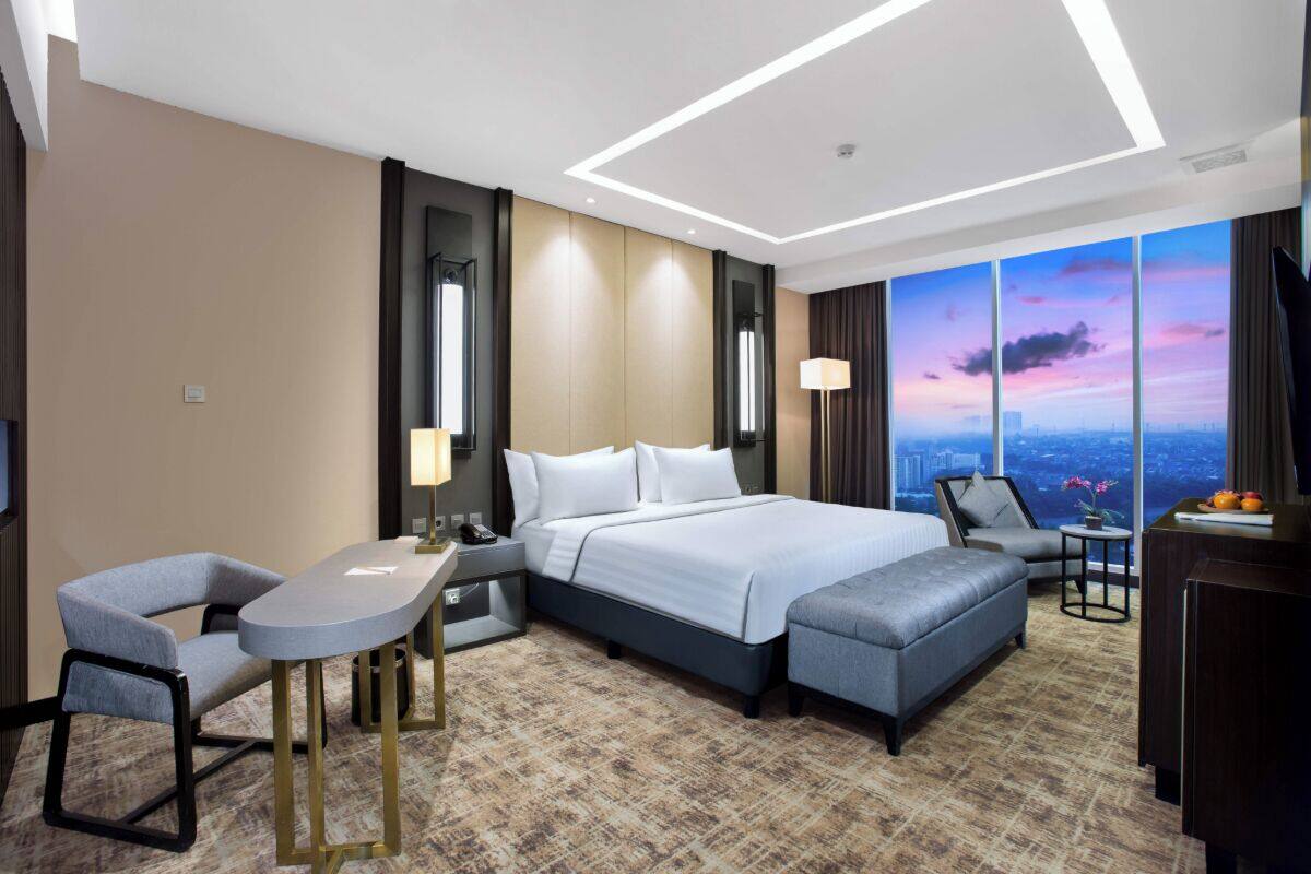 A modern hotel room with a large bed, a desk, a chair, and a view of the cityscape.