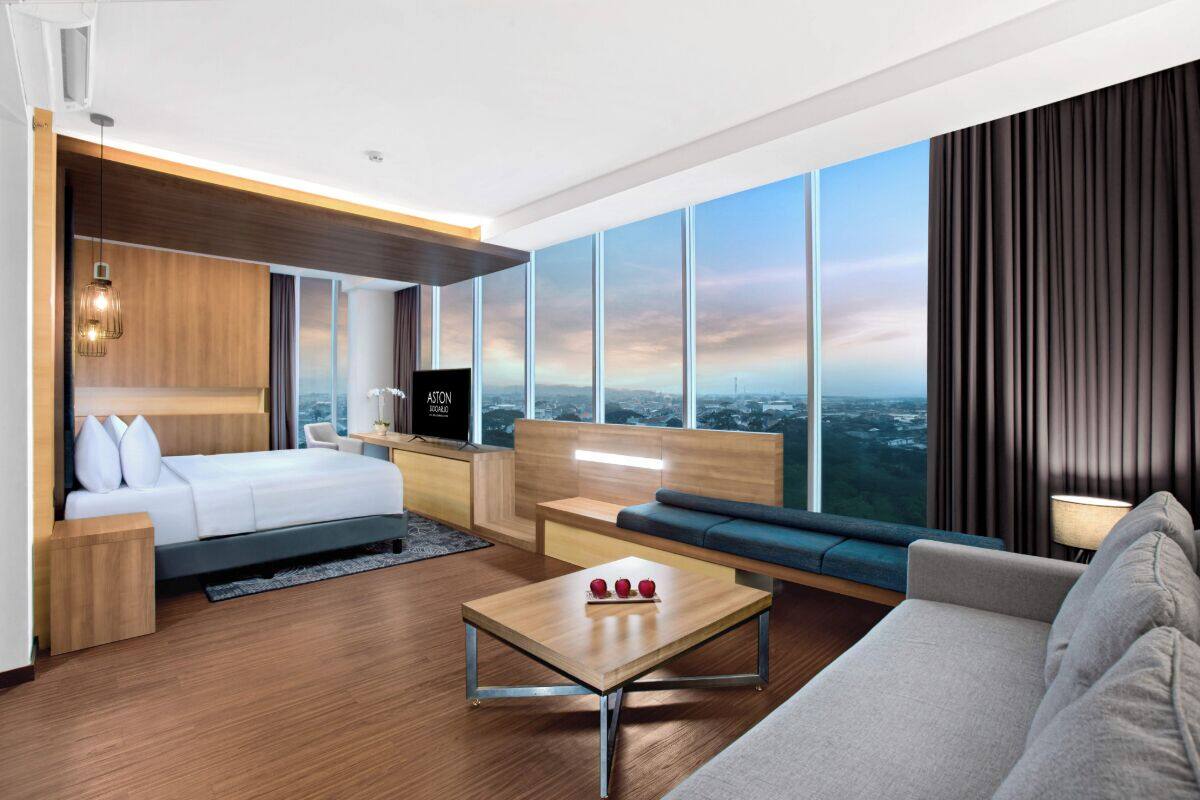 A modern hotel room with a large bed, a sofa, a coffee table, and a TV. The room has large windows with a city view.