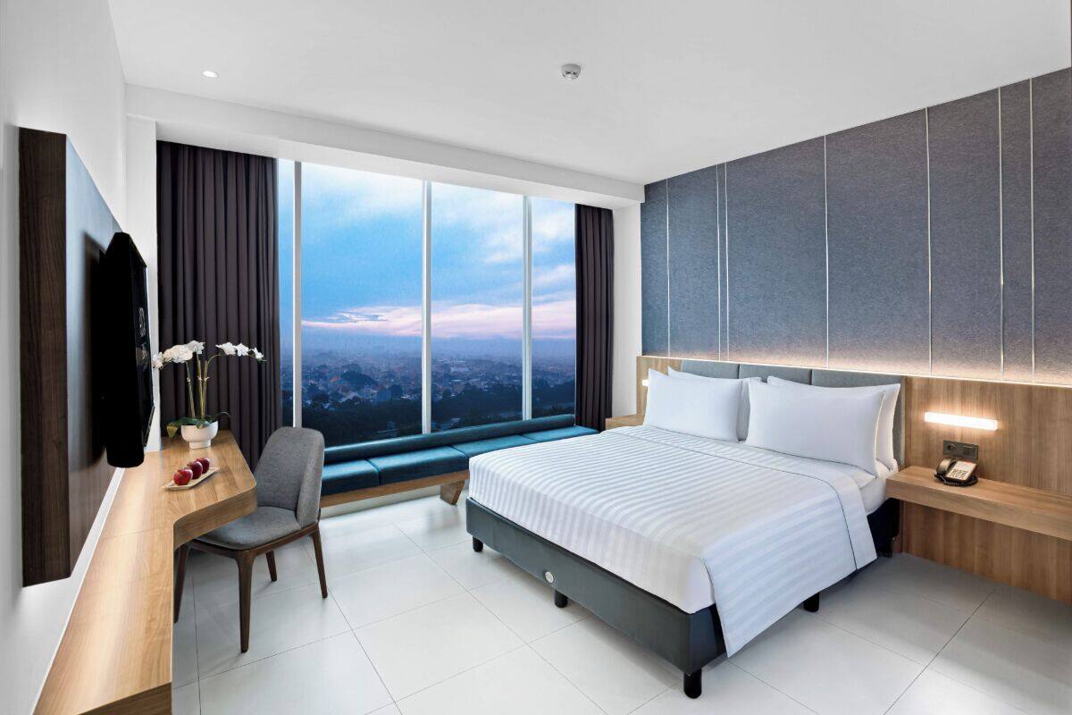 A modern hotel room with a large bed, a desk with a chair, a TV, and a view of the city through large windows.