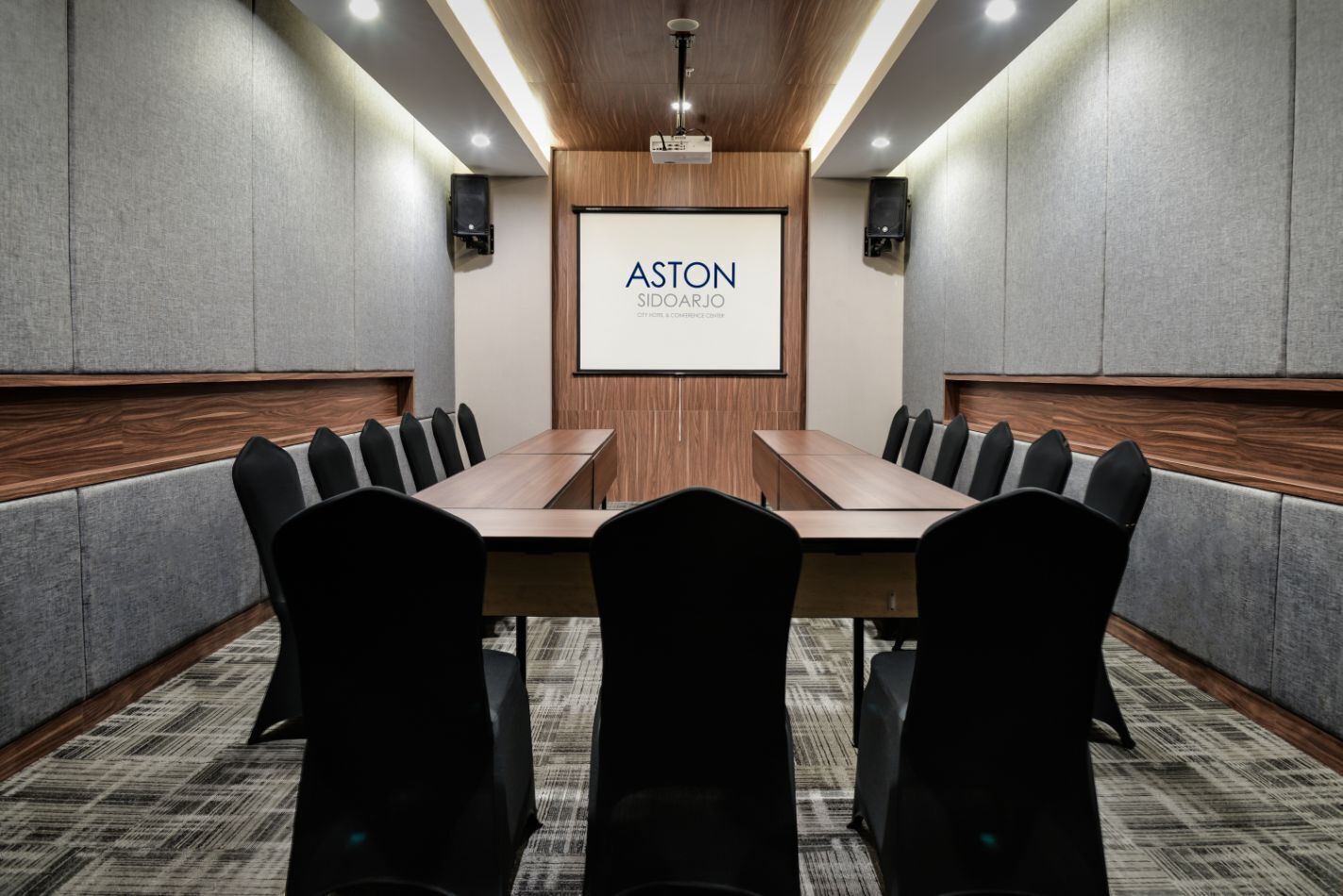 A conference room with a long table, black chairs, a projector screen, and wooden paneling.