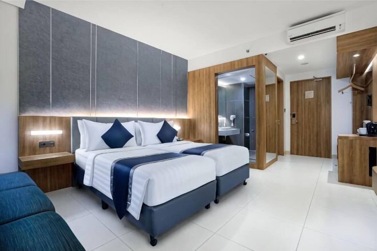 A modern hotel room with two beds, a wooden wardrobe, a mirror, and a small kitchenette.