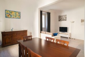 A dining area with a wooden table and chairs, a wooden cabinet, a painting on the wall, a television on a white stand, and a floor lamp.
