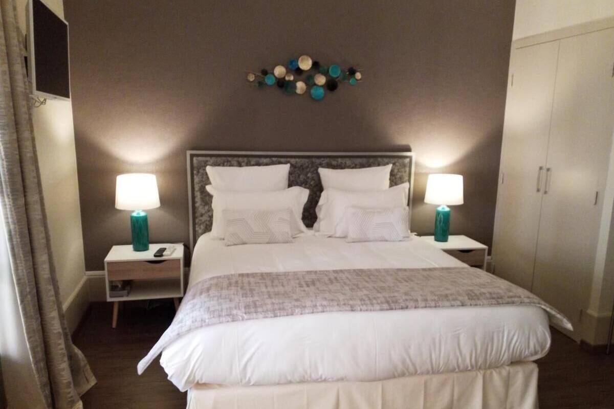 Uma cama bem arrumada com lençóis brancos e um cobertor cinza, ladeada por duas mesas de cabeceira com abajures verde-água, uma peça decorativa na parede e um guarda-roupa.