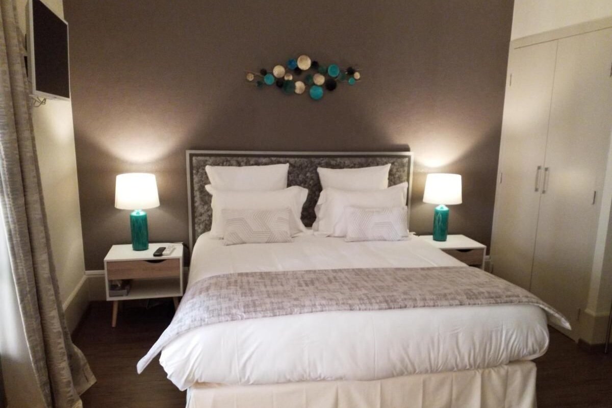 A neatly made bed with white linens and a gray blanket, flanked by two bedside tables with teal lamps, a wall-mounted decorative piece, and a wardrobe.