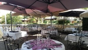 Un área de comedor al aire libre con mesas redondas, cada una con mantel blanco, servilletas moradas y copas de vino.