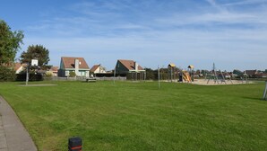 Un parque con césped, juegos infantiles, casas al fondo y cielo despejado.