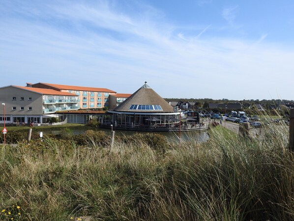 A waterfront building with a thatched roof and a glass extension, surrounded by a grassy area and a parking lot.