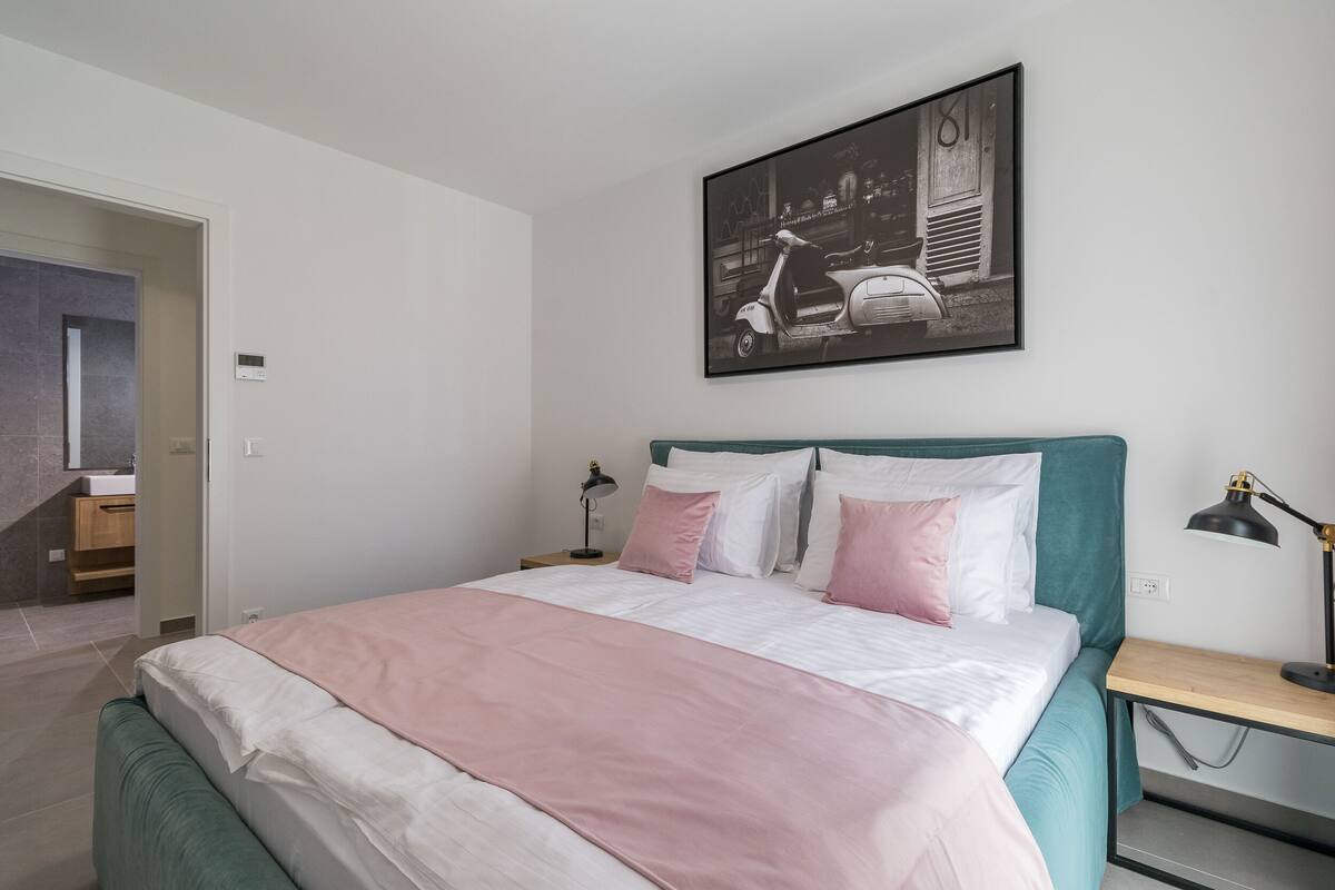 A neatly arranged bedroom with a bed, bedside tables, a wall-mounted picture, and a door leading to a bathroom.