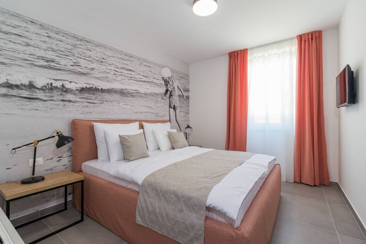 A bedroom with a bed, a nightstand, a lamp, a wall with a textured design, a window with curtains, and a TV mounted on the wall.