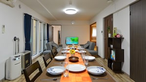 Un comedor con una mesa larga dispuesta para seis personas, un televisor montado en la pared y una estantería con libros y objetos decorativos.