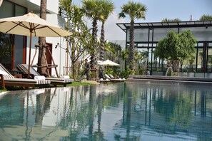 Zona de piscina con sillas de descanso y sombrillas, rodeada de palmeras y arquitectura moderna.