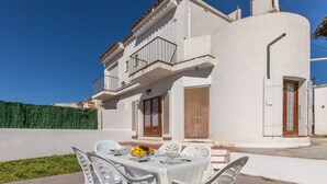 Una villa blanca con balcón, un comedor con sillas de plástico y una mesa preparada para cuatro, y un seto verde al fondo.