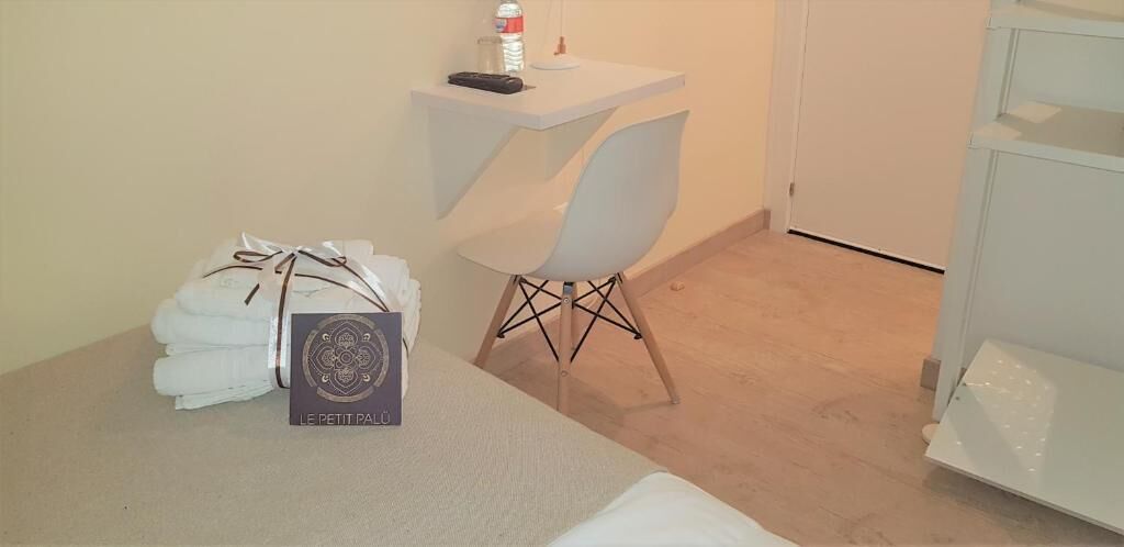 A neatly arranged room with a bed, a small table with a remote control and a bottle, a chair, and a stack of folded towels.