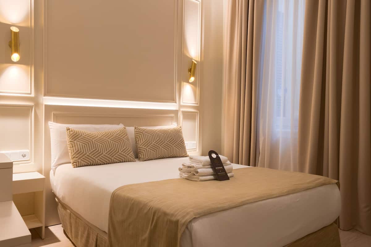 A neatly made bed with a beige bedspread and pillows, a bedside table with a lamp, and a window with curtains.