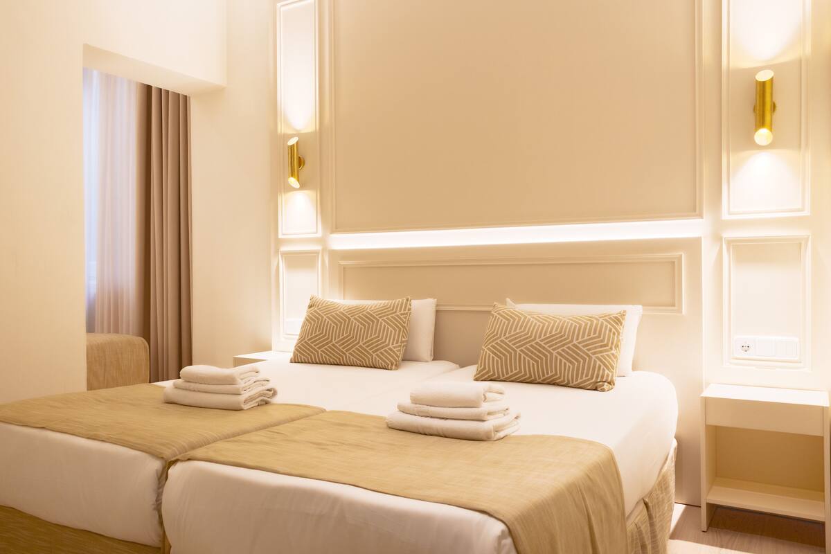 A hotel room with two beds, a bedside table, a nightstand, and a window with curtains.