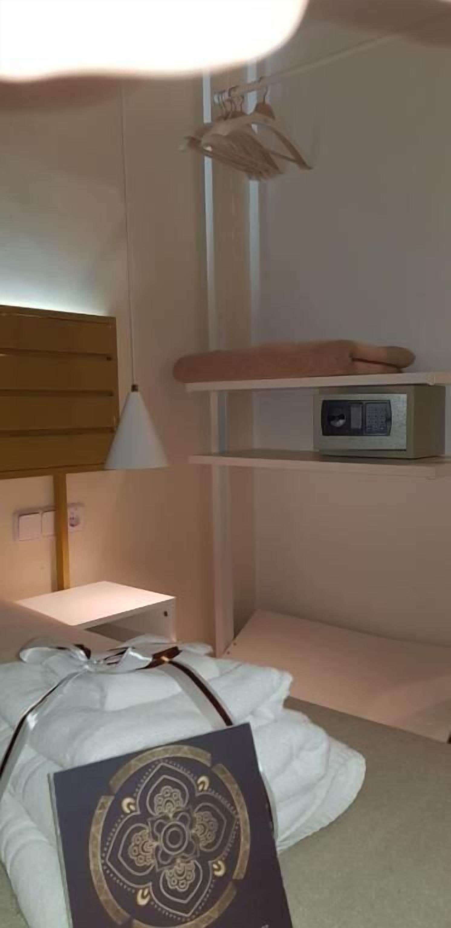 A bedroom with a bed, a bedside table, a lamp, a shelf with a radio, and a hanging light fixture.