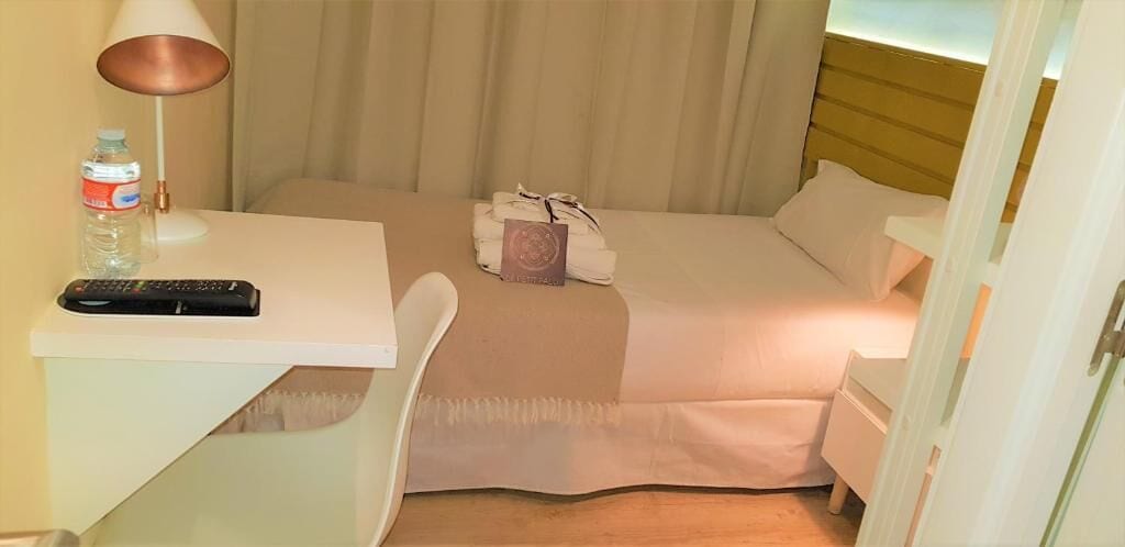 Ein Hotelzimmer mit einem Bett, einem Nachttisch mit Fernbedienung, einer Lampe und einer Wasserflasche.
