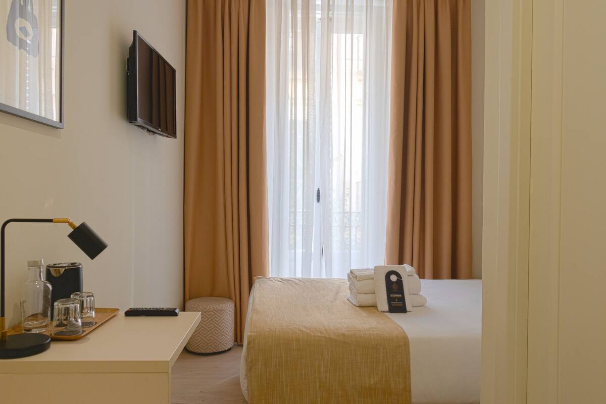 A hotel room with a bed, a bedside table with a lamp, a remote control, and a tray with glasses and a bottle.