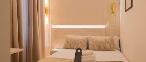 A neatly made bed with patterned pillows, a folded towel, and a room with a window and curtains.