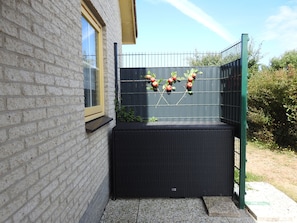 Un coin d’un bâtiment avec un mur en briques, une fenêtre et un espace extérieur aménagé avec des sièges noirs, une clôture en métal et des plantes.
