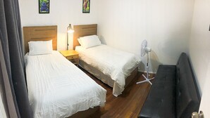 Et værelse med to enkeltsenge, et natbord med lampe, en gulvventilator og en sort bænk.
