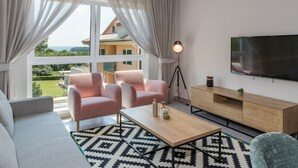 Ein modernes Wohnzimmer mit einer Couch, zwei Sesseln, einem Couchtisch und einem Fernseher. Große Fenster bieten Blick auf ein Gebäude und Grünflächen.