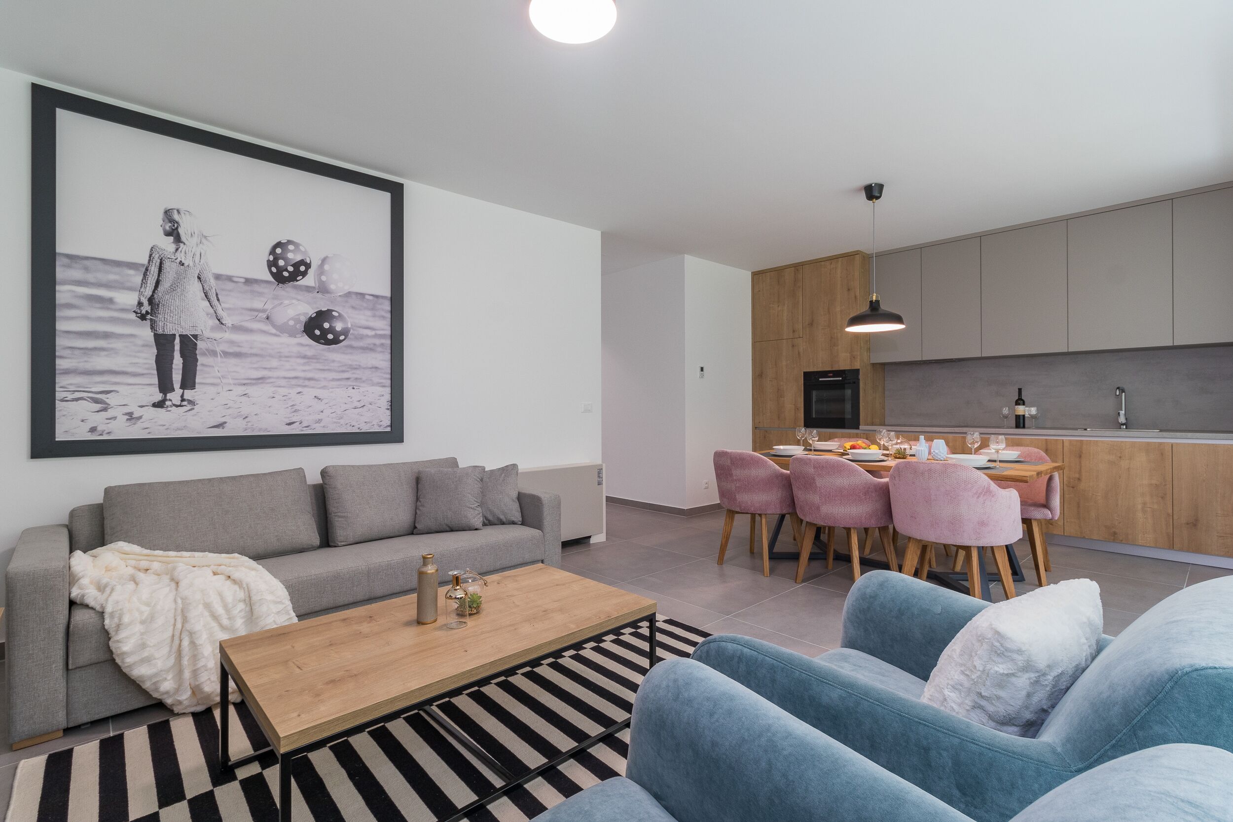 A modern living room with a grey sofa, a blue sofa, a wooden coffee table, and a black and white striped rug. There is a dining area with pink chairs and a wooden table.