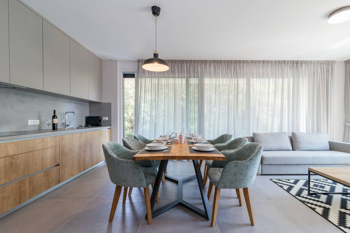 Een moderne keuken met houten kastjes, een eettafel gedekt voor zes personen en een groot raam met dunne gordijnen.