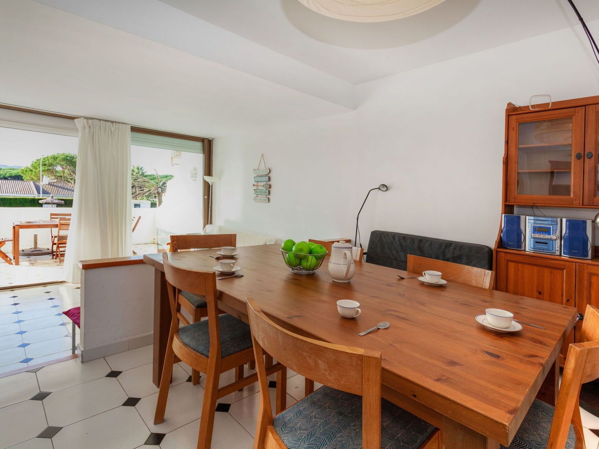 A dining area with a wooden table, chairs, a bowl of green apples, a white kettle, and a cabinet with electronic devices.