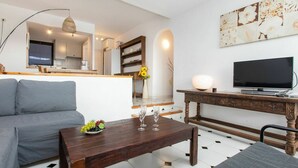 A modern living room with a grey sofa, a wooden coffee table with a fruit bowl and wine glasses, a TV on a wooden stand, and a kitchen in the background. - Nice apartment for 4 guests with WIFI, pool, TV and terrace (Torroella De Montgrí)