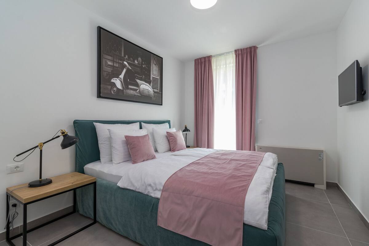 Quarto com cama, mesa de cabeceira com luminária, televisão na parede e janela com cortinas.