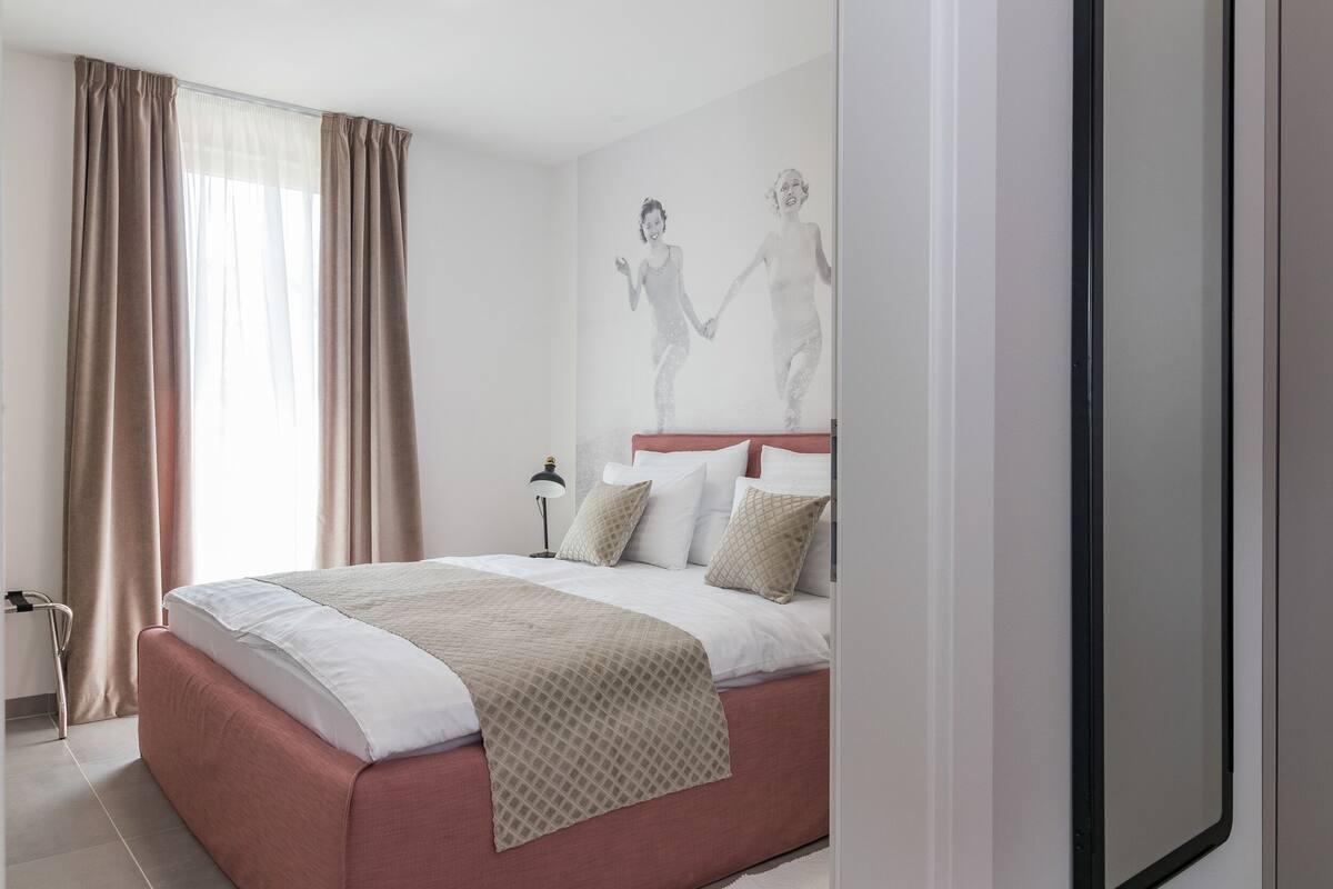 Cama bem arrumada com manta estampada, abajur e parede com arte minimalista.
