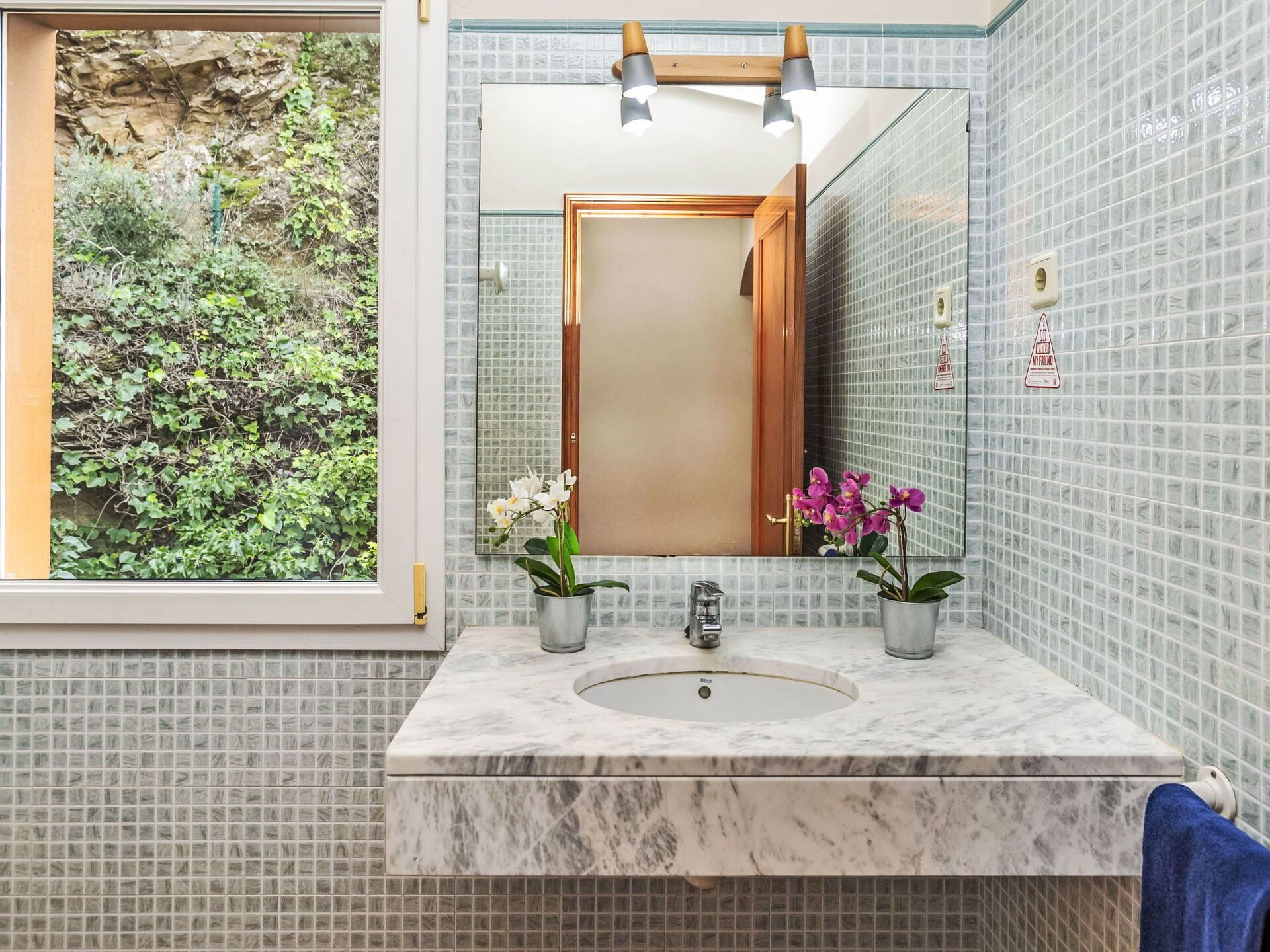A bathroom with a marble sink, two potted orchids, a mirror, and a view of greenery outside.