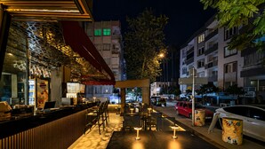 Un área de comedor al aire libre con mesas y sillas, una barra y vista a la calle y edificios de fondo.