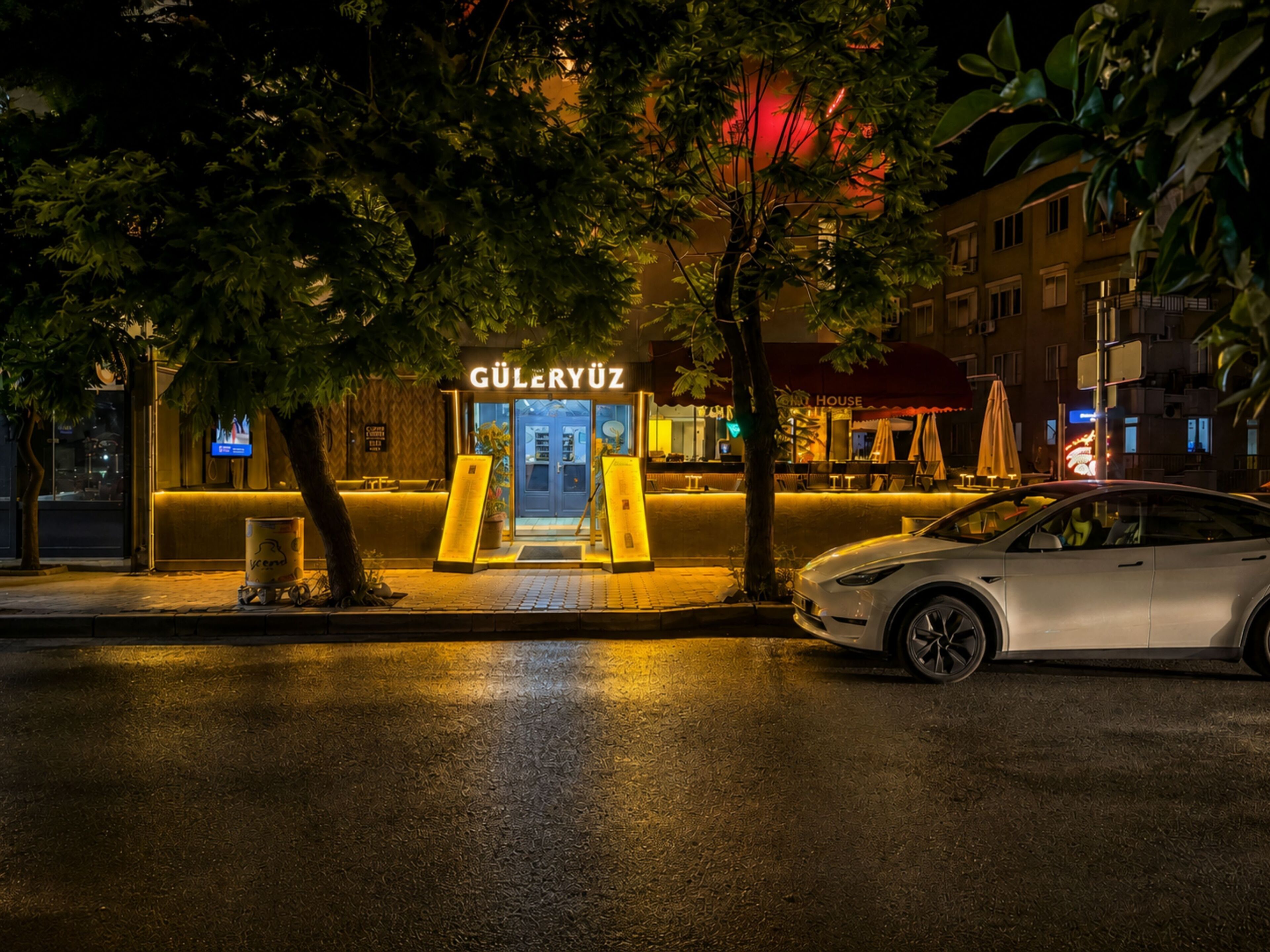 A building with a sign reading 'GÜLERYUZ' at night, a car parked in front, and trees in the foreground.