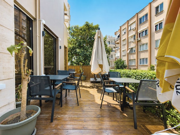 Un espacio de patio al aire libre con mesas y sillas, rodeado por una terraza de madera, y contiguo a un edificio con varias ventanas.