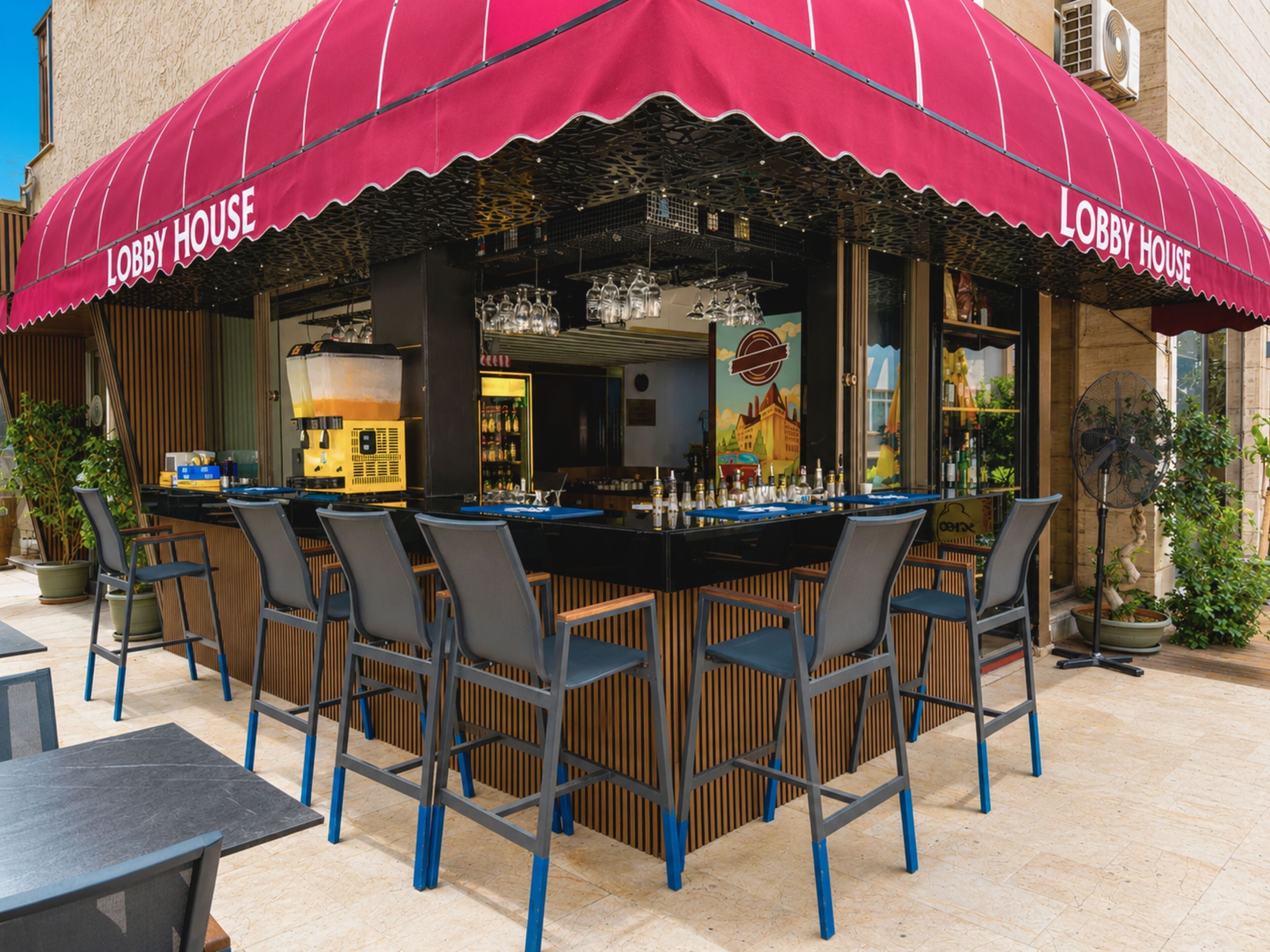 An outdoor bar area with a red awning labeled 'Lobby House,' featuring a bar counter, multiple chairs, and a fan.