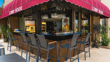 An outdoor bar area with a red awning labeled 'Lobby House,' featuring a bar counter, multiple chairs, and a fan.