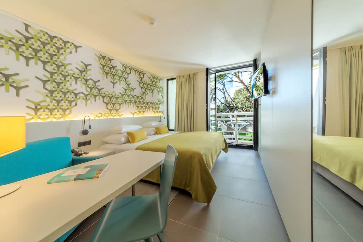 A hotel room with a bed, a desk, a chair, a TV, and a balcony with a view of the outside.