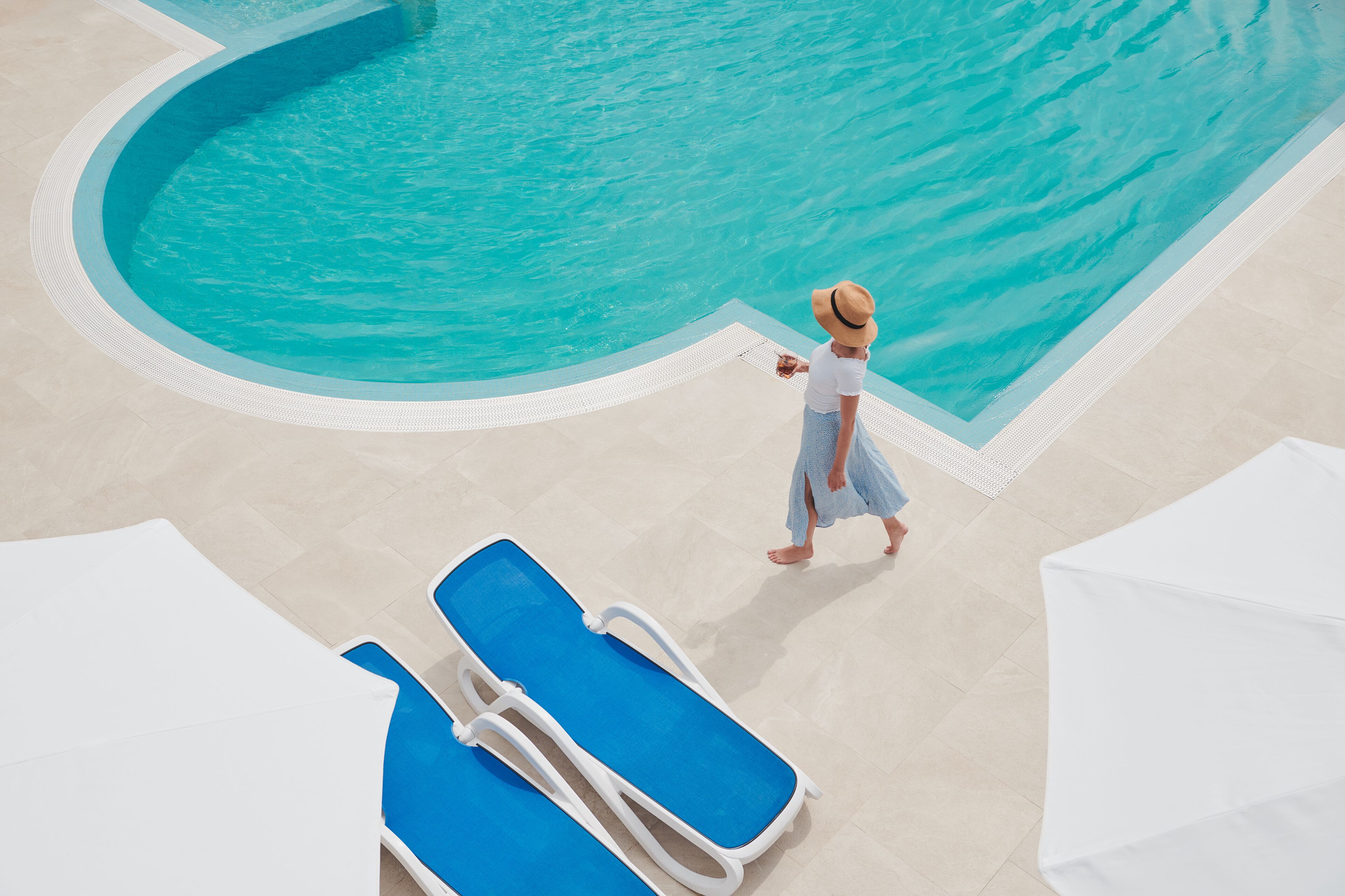 A pool area with a person walking by, lounge chairs, and a tiled floor.