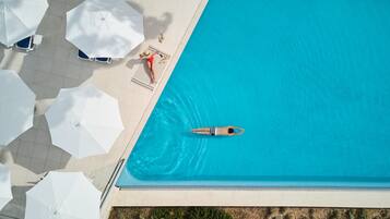 Vue aérienne d’une piscine à l’eau d’un bleu limpide, entourée de parasols blancs et de chaises longues.