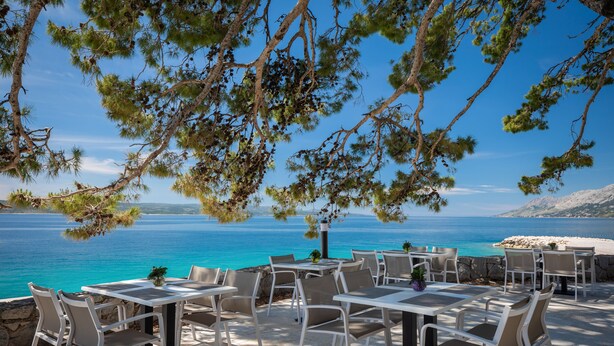 An outdoor seating area with tables and chairs overlooking a beach and the sea. - Bluesun Mala Berulia (Brela)