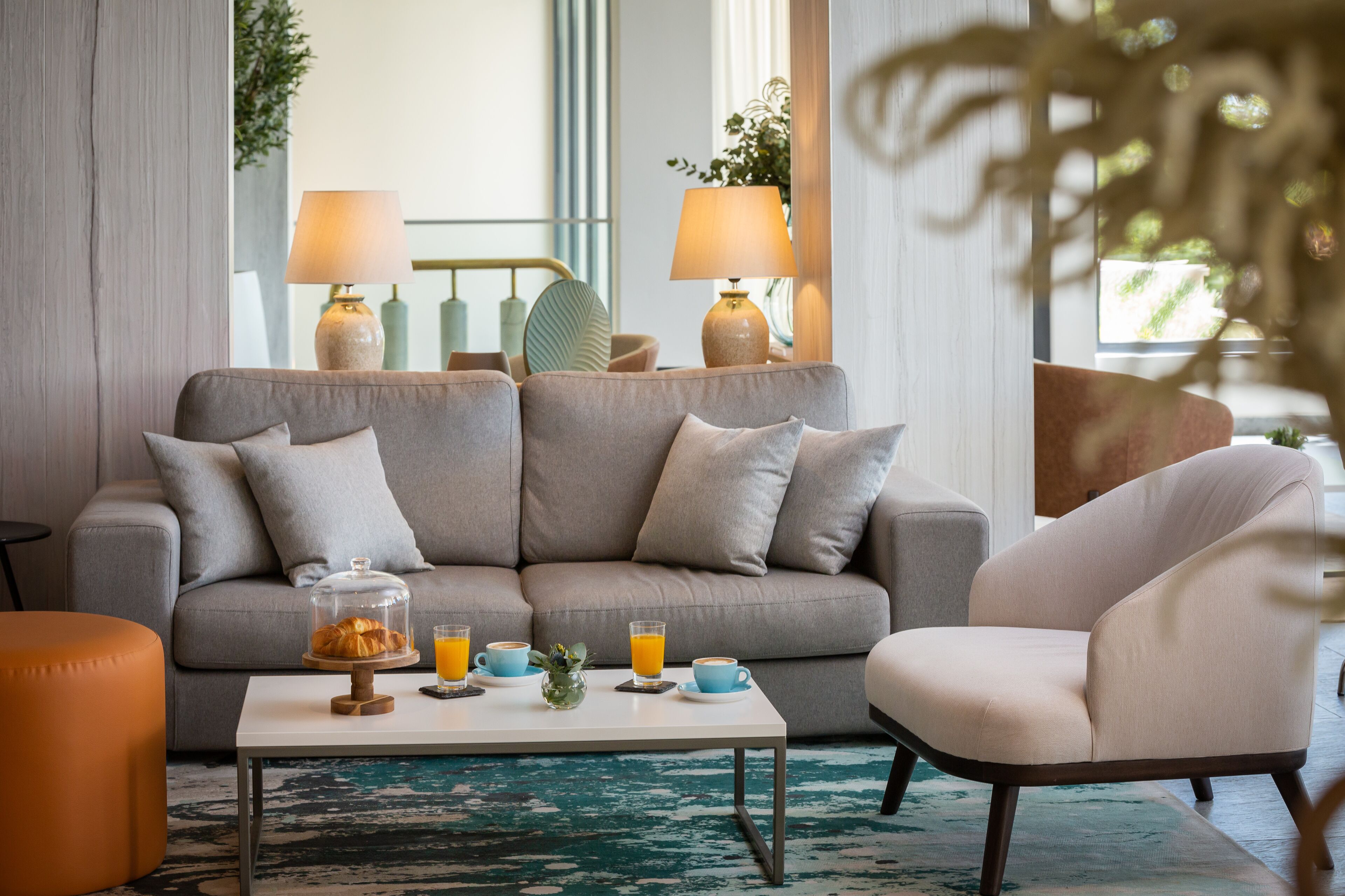 A modern living room with a grey sofa, a white coffee table, and a cream armchair. There are two lamps on the sofa and a glass coffee pot on the table.
