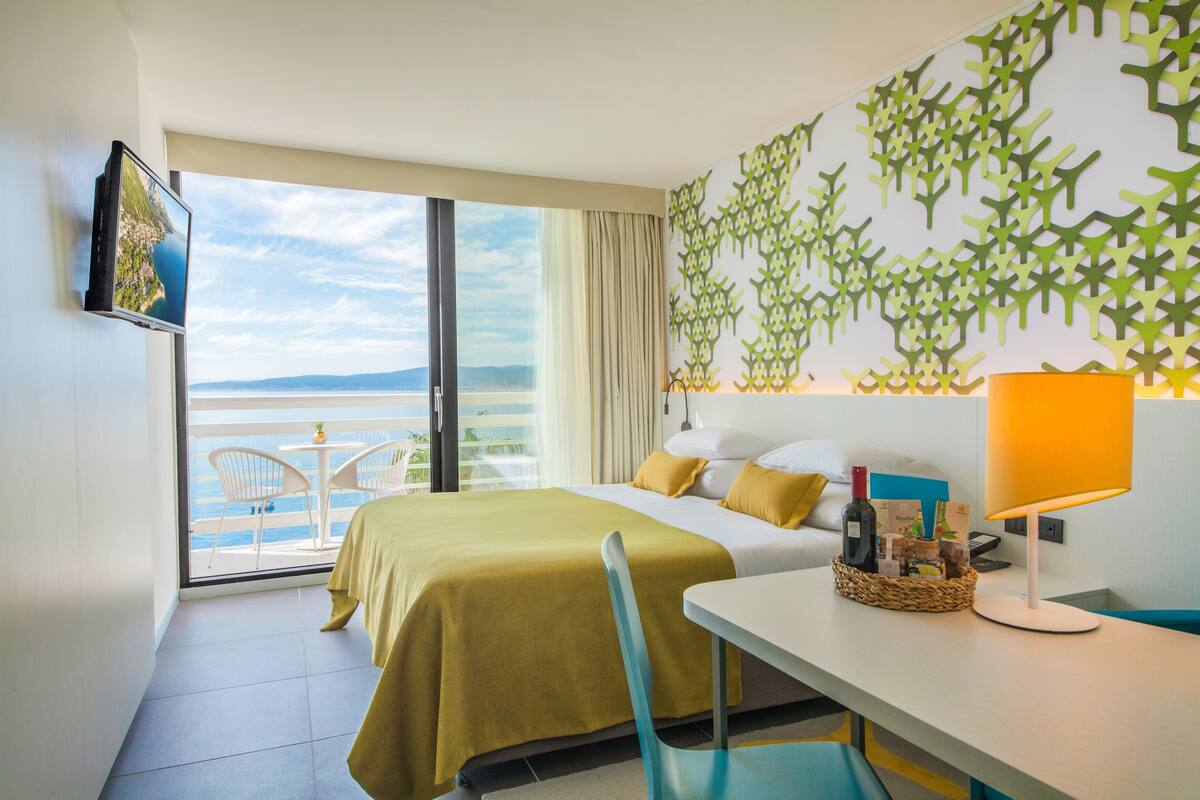 A hotel room with a bed, a desk, a lamp, a TV, and a view of the sea.