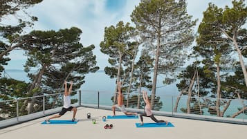 Deux personnes pratiquent le yoga sur des tapis bleus, avec des kettlebells et des haltères, sur un balcon d’où l’on peut admirer des arbres et l’eau.