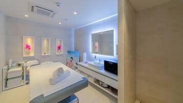 A modern hotel room with a bed, a vanity with a mirror, a sink, and a massage table.